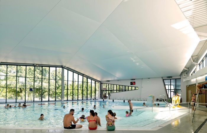 Intérieur du centre aquatique A26 baigné de lumière naturelle grâce à de grandes baies vitrées, avec des résidents profitant des installations aquatiques, mettant en lumière un espace convivial et énergétiquement efficace