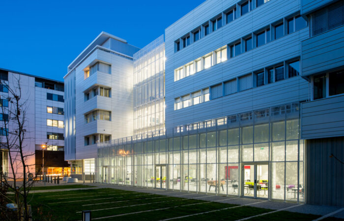Vue du crépuscule sur le campus moderne de l'agence d'architecture A26, mettant en évidence les façades en verre et métal, l'éclairage intérieur chaleureux et les espaces verts soigneusement aménagés.